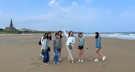 Seven boarders from Harrogate Ladies' College stand on the beach at Tynemouth, under blue skies, with the sea next to them