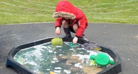 A boy from Highfield Pre-School, wearing a red coat with the hood up, bends over to play with toys in a sandpit filled with water during wet weather